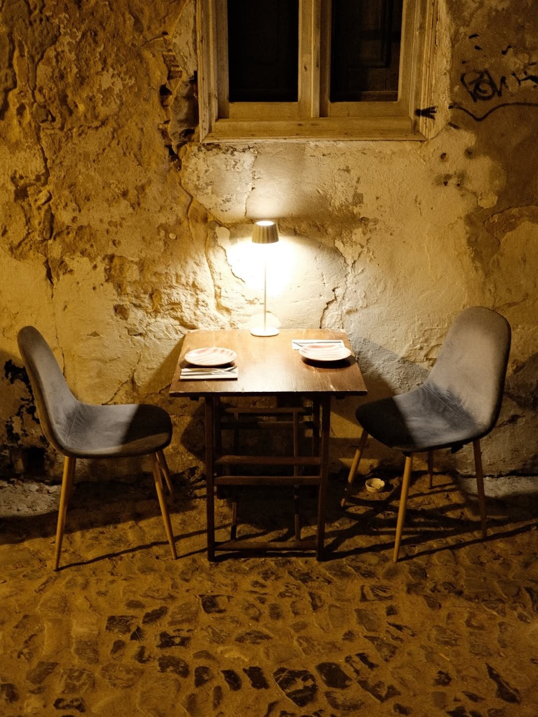 Mesa para dois no beco empedrado, iluminada por um pequeno candeeiro junto a uma parede antiga