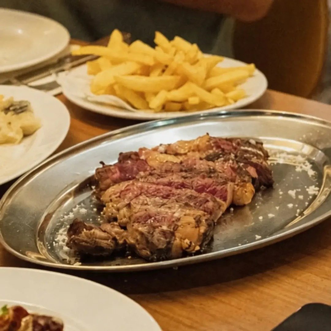 Carne grelhada fatiada numa travessa de inox, com batatas fritas ao lado